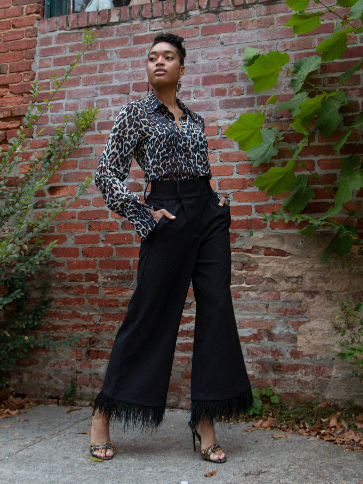 Person wearing a leopard print blouse and black pants standing against a brick wall.